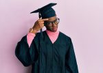 Young african american girl wearing graduation cap and ceremony robe pointing unhappy to pimple on forehead, ugly infection of blackhead. acne and skin problem