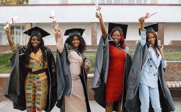 Excited african american female students at their graduation. Group of young afro american female student dressed in black graduation gown. Campus as a background. Girls cheerfully smiling with arms up, holding diploma.