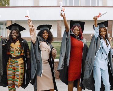 Excited african american female students at their graduation. Group of young afro american female student dressed in black graduation gown. Campus as a background. Girls cheerfully smiling with arms up, holding diploma.