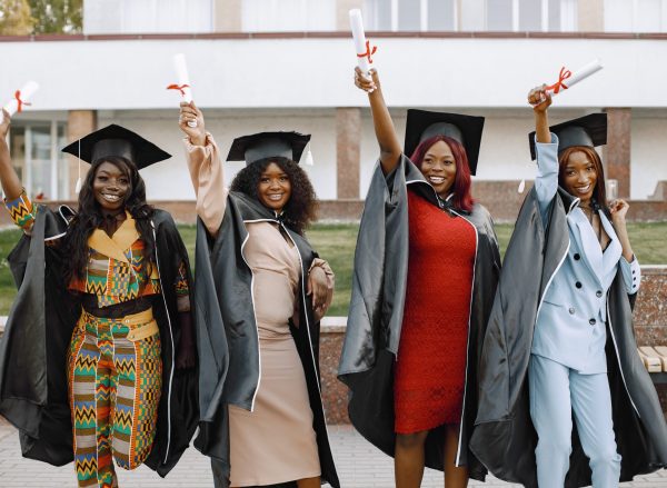 Excited african american female students at their graduation. Group of young afro american female student dressed in black graduation gown. Campus as a background. Girls cheerfully smiling with arms up, holding diploma.