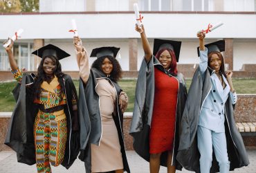 Group of young afro american female student dressed in black graduation gown. Campus as a background. Girls cheerfully smiling with arms up, holding diploma.