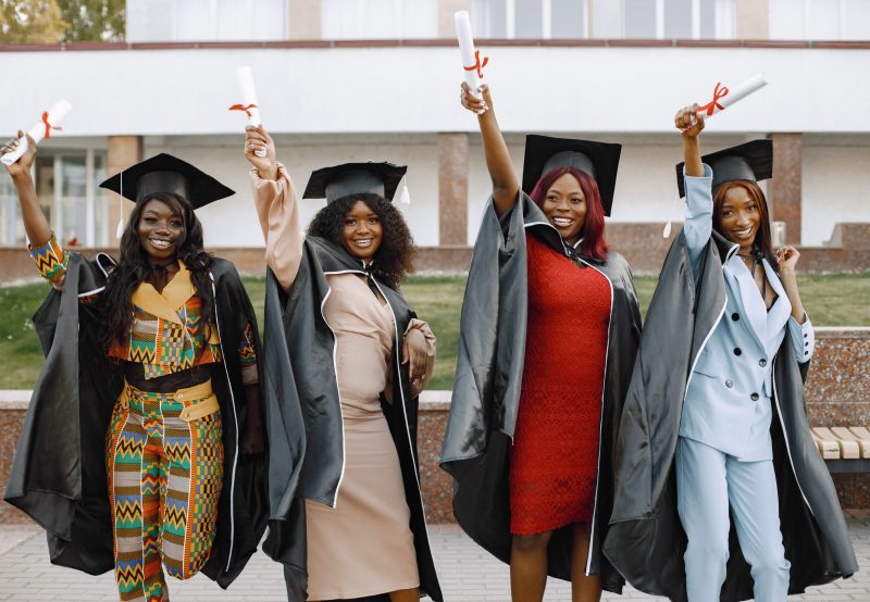 Group of young afro american female student dressed in black graduation gown. Campus as a background. Girls cheerfully smiling with arms up, holding diploma.