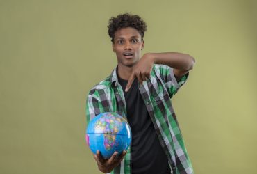 young african american traveler man holding globe pointing with finger to it looking at camera exited standing over green background