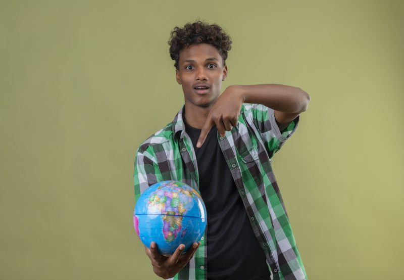 young african american traveler man holding globe pointing with finger to it looking at camera exited standing over green background